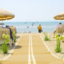 Photograph of Mobi- Path / Roadway running along sand down towards the sea and a beach with beach umbrellas and chairs either side of the path. There are green and yellow plants either side of the path and people and boats in the sea.
