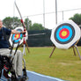 Photograph of a disabled archer on a blue Mobi- Path / Roadway that has been rolled out on grass, shooting an arrow at a bright target