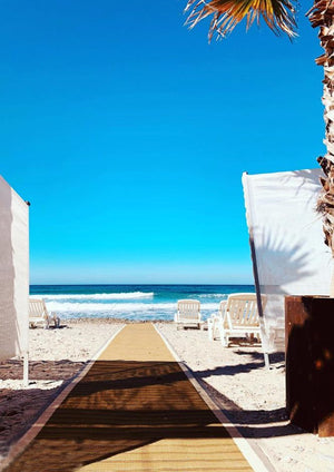Photograph of bright blue sky with a brown Mobi-Path / Roadway running along sand on a beach towards the sea. With beach chairs and waves in the sea.