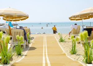 Photograph of Mobi- Path / Roadway running along sand down towards the sea and a beach with beach umbrellas and chairs either side of the path. There are green and yellow plants either side of the path and people and boats in the sea.  