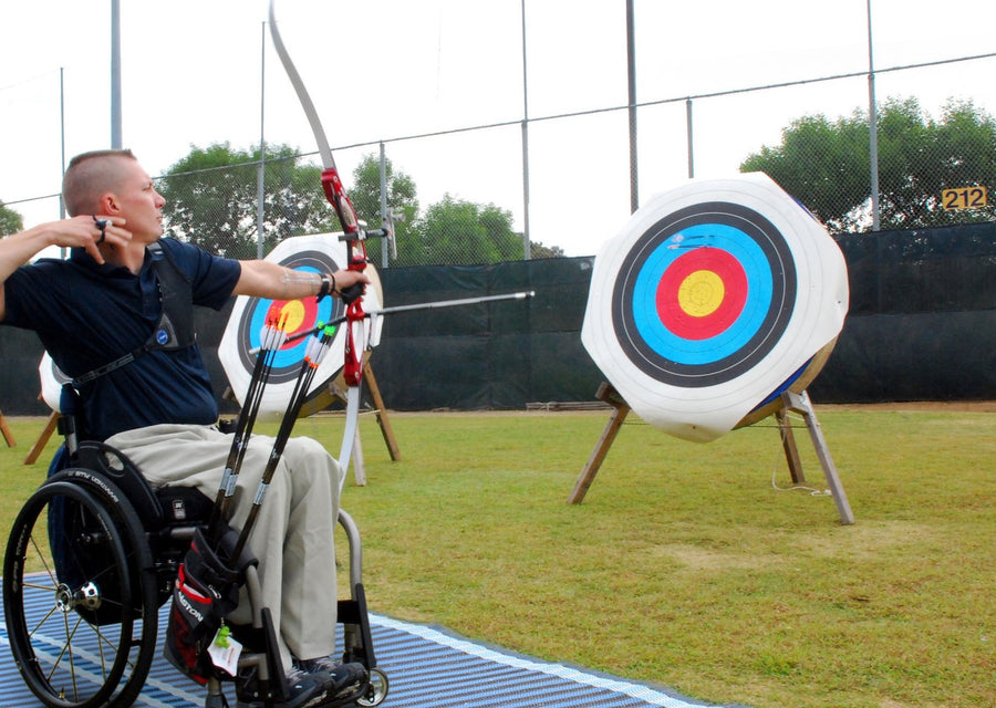 Photograph of a disabled archer on a blue Mobi- Path / Roadway that has been rolled out on grass, shooting an arrow at a bright target 