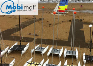 There are 10 catamarans with no sails  in the foreground on a beach. Past the catamarans are three Mobi-Boat Ramps intersecting with each other to form pathways. There are two catamarans in the distance with their sails up and there is water past those boats. 