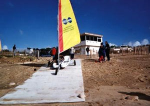 A Mobi-Boat Ramp has been rolled out on a sandy and slightly rocky surface from a sailing clubhouse presumably down to water. A sailing boat with a bright yellow and red sail on a trailer in being wheeled along the ramp while a couple people watch. There is a bright blue sky in the background. 