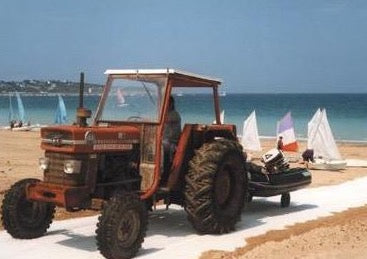 A Mobi-Boat Ramp has been rolled out on sand on a beach. An old red tractor has pulled a dingy out of the water and is pulling it along the boat ramp
