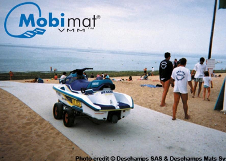 Mobi-Boat Ramp has been rolled out on sand. A jet ski on a trailer is on the ramp. There is water in the distance and lots of people in the photo. 