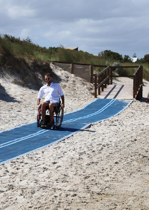Photograph of an Australian beach with a person in a wheelchair moving along a blue Mobi-Mat that has extended access along the beach.   Mobi-Fence in the background, running along the beach access path.