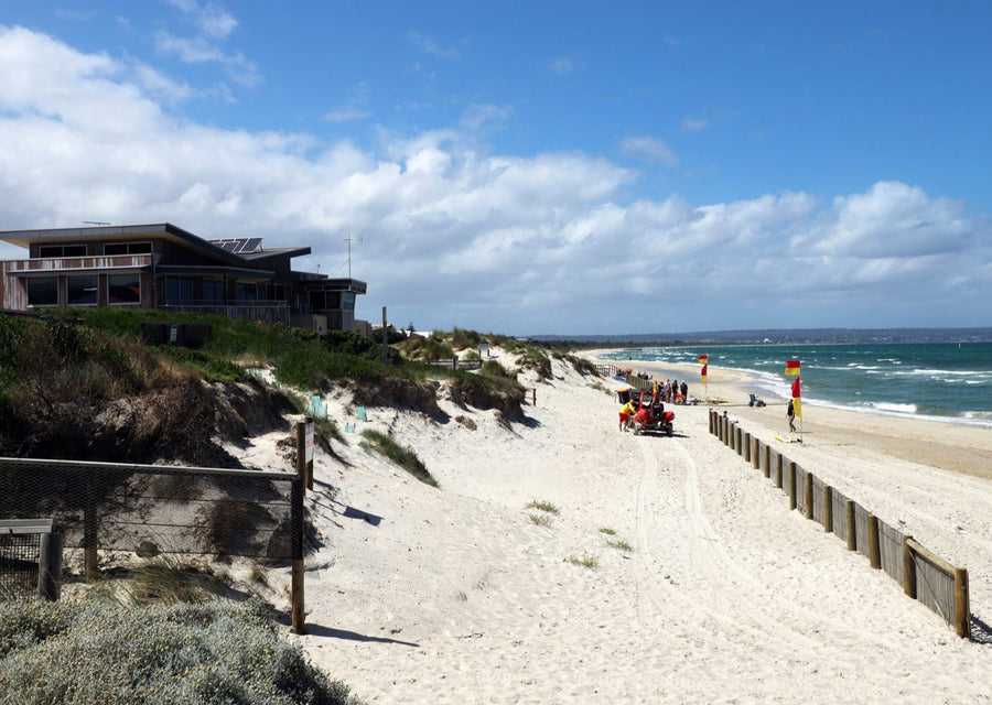 Photo of Surf Club building and beach view. Surf-lifesaving flags are set up on the beach with Surf-Lifesavers in the distance. Mobi-Fence is in the foreground running horizontally with the coastline and the ocean to help stop sand movement. 