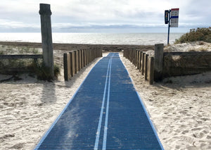 Photograph of an Australian beach that has a blue Mobi-Mat running down to the sea with a Mobi-Fence running along each side of the Mobi-Mat. Sea in the distance. Sand in the foreground.  
