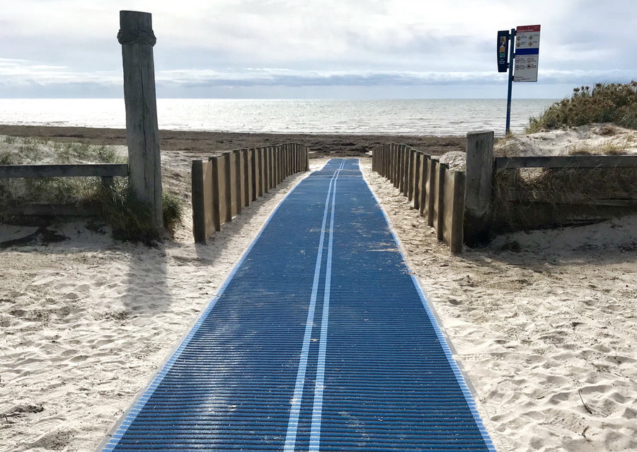 Photograph of an Australian beach that has a blue Mobi-Mat running down to the sea with a Mobi-Fence running along each side of the Mobi-Mat. Sea in the distance. Sand in the foreground.  