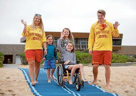 Mob-Path being used on the beach by a child in a three wheeled wheelchair with two surf lifesavers and two other children