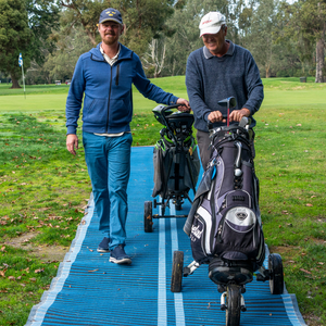 Photograph of two men wearing caps, one pulling a golf buggy and the other is pushing a golf buggy on a blue Mobi-Path rolled out on grass.
