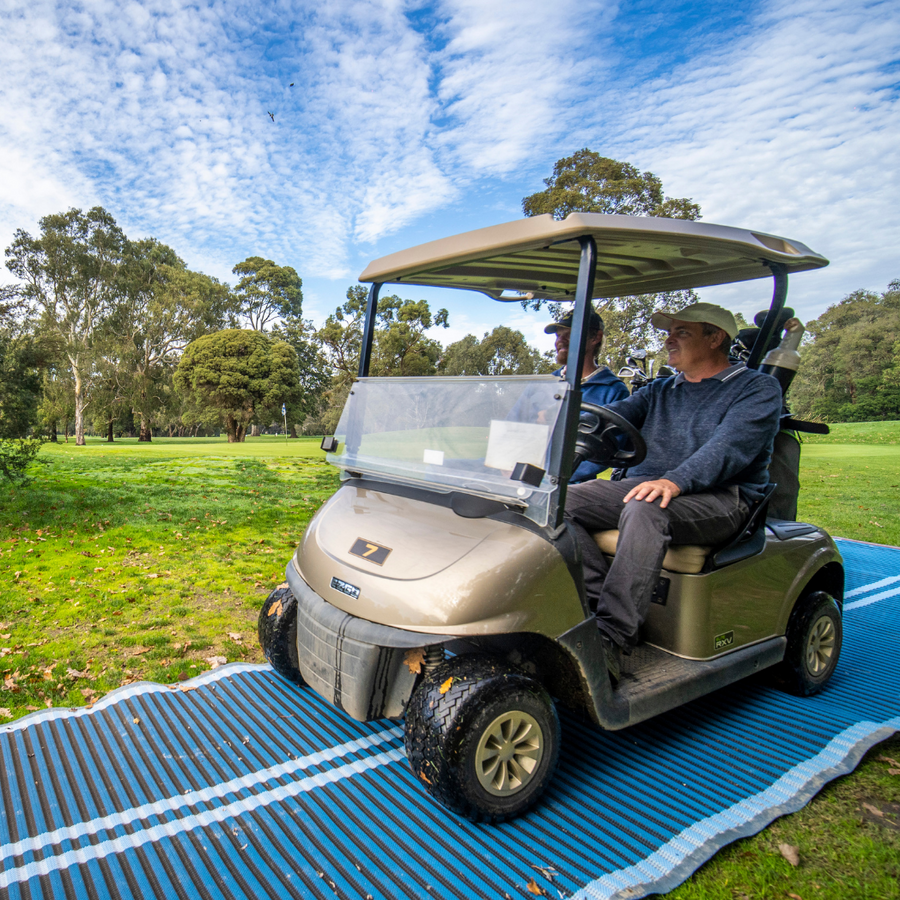 Photograph of two men wearing caps riding a golf cart on a blue rolled out Mobi-Path which is on grass.