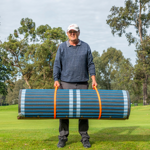 Photograph of man in cap standing on grass and lifting a blue rolled up mobi mat