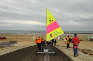Sailing boat with bright yellow and pink sail being rolled on wheels along a Mobi-Boat Ramp mat that has been rolled out on sand on a long beach