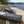 A brown Mobi-Boat Ramp with clear white lines has been rolled out next to a wooden boat ramp on the edge of a lake. There are two aluminium rowing boats on the ramp. The lake  is in the background with trees growing the lake edge.