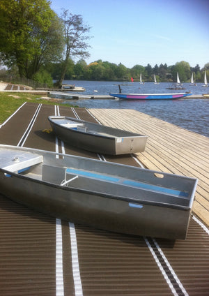 A brown Mobi-Boat Ramp with clear white lines has been rolled out next to a wooden boat ramp on the edge of a lake. There are two aluminium rowing boats on the ramp. The lake  is in the background with trees growing the lake edge.