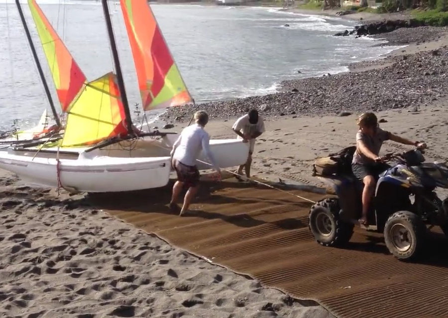 A brown Mobi-Boat Ramp has been rolled out on sand on a sandy and rocky beach. A catamaran is attached to a quad bike by a rope and is being pulled up the ramp. Two additional people are assisting to guide the direction of the catamaran. 
