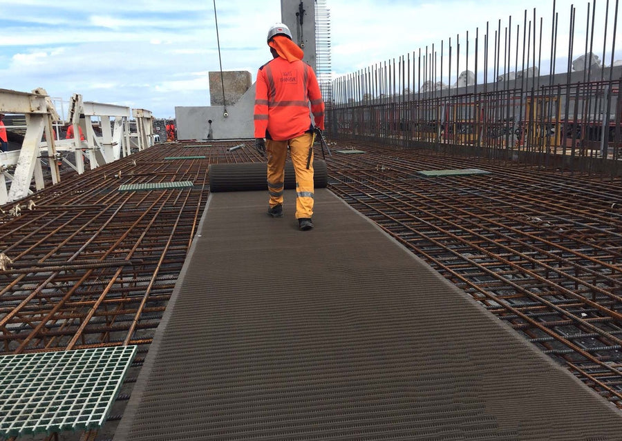 Photograph of a workman rolling out a Mobi- Path / Roadway along reinforced iron mesh at a construction site. Walking away from photographer. 