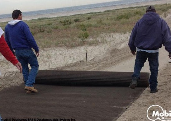 Two people rolling out a Mobi-Boat Ramp along a sandy beach track towards the ocean