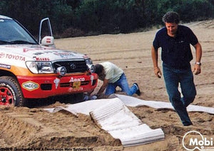 Photo of a bogged 4WD on rugged sandy ground with two men setting up white Mobi-Tracks under the cars wheels.